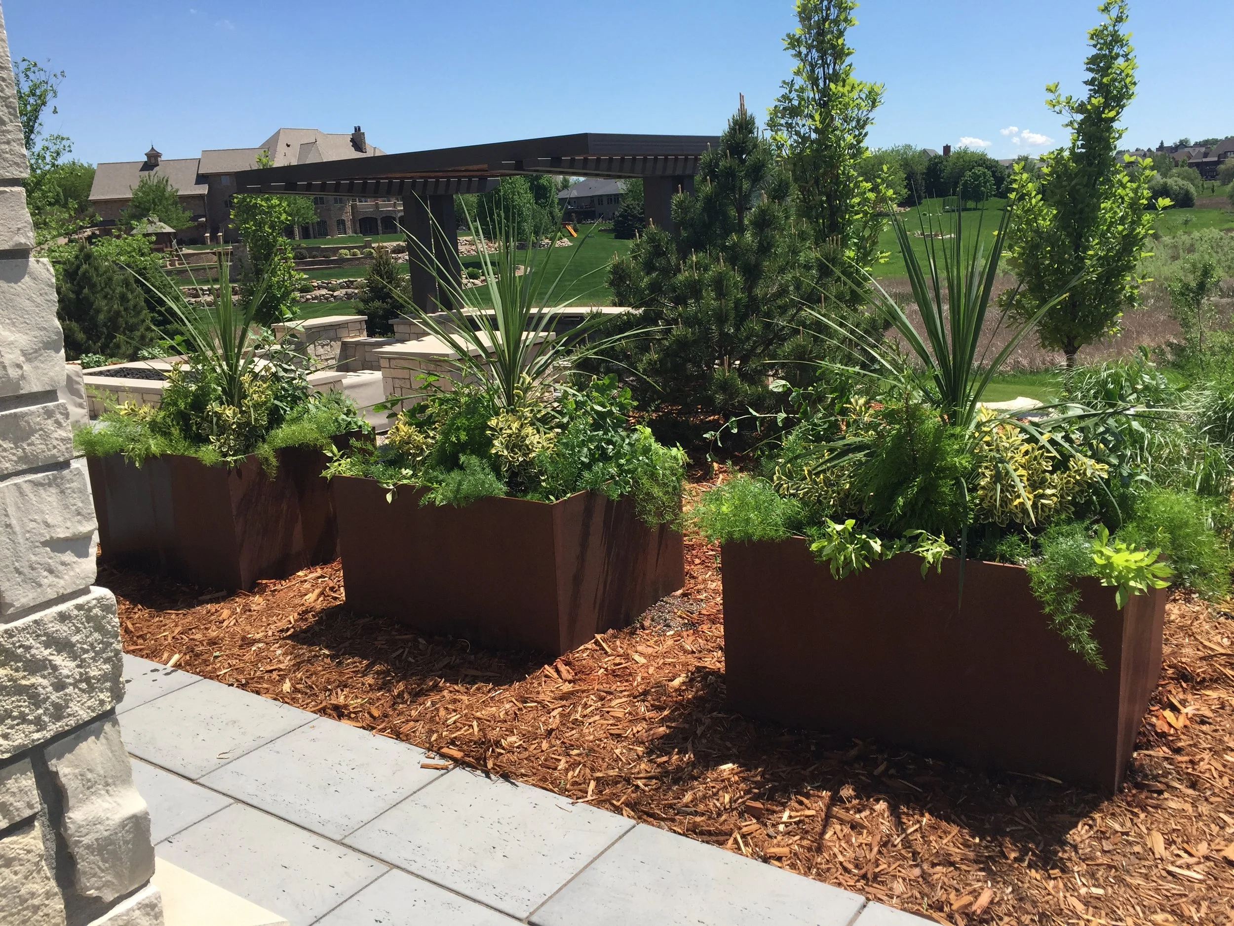 Three rust-colored rectangular planters filled with green plants, set in a landscaped area with mulch and a patio, with trees and a house in the background.