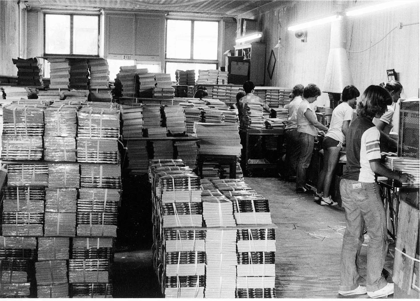 A room filled with stacks of papers and magazines, with people standing at desks working on them in an indoor office setting.