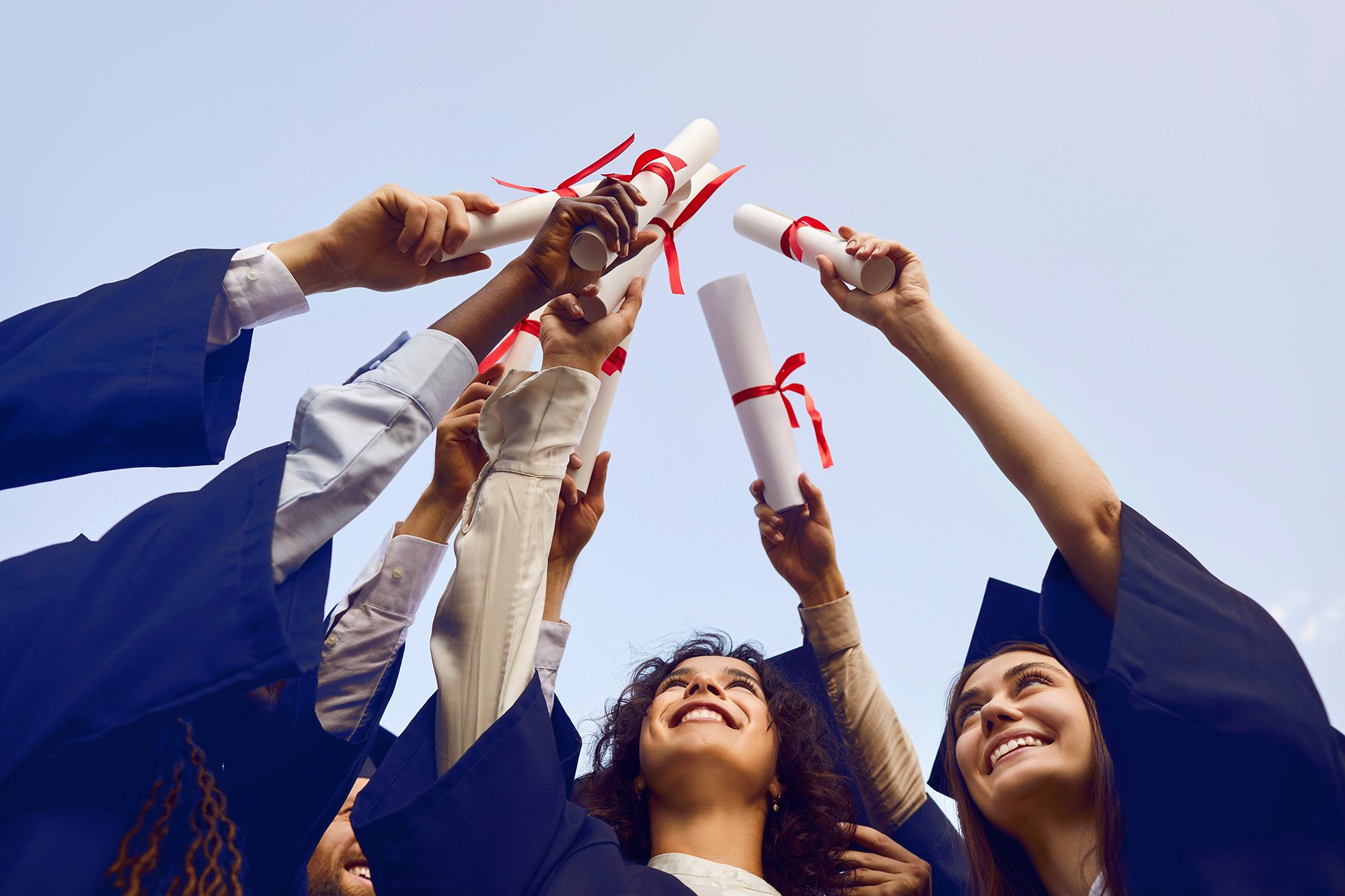 Group of graduates in caps and gowns celebrating, holding diplomas tied with red ribbons up towards the sky.