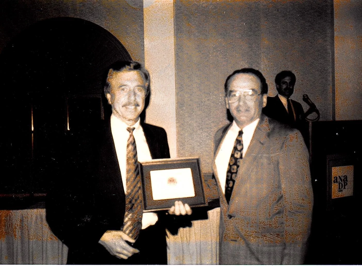 Two men in suits standing indoors, one presenting a framed certificate or award to the other. In the background, a man is at a podium with a microphone, standing next to a sign that reads "Ayan DP."