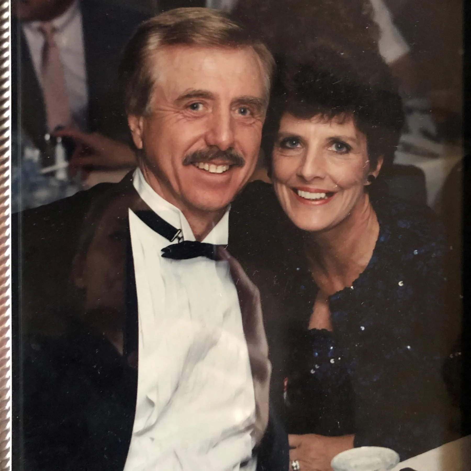 A smiling man with a mustache in a tuxedo and a woman with short dark hair in a sparkly dress, sitting closely together at a formal event.