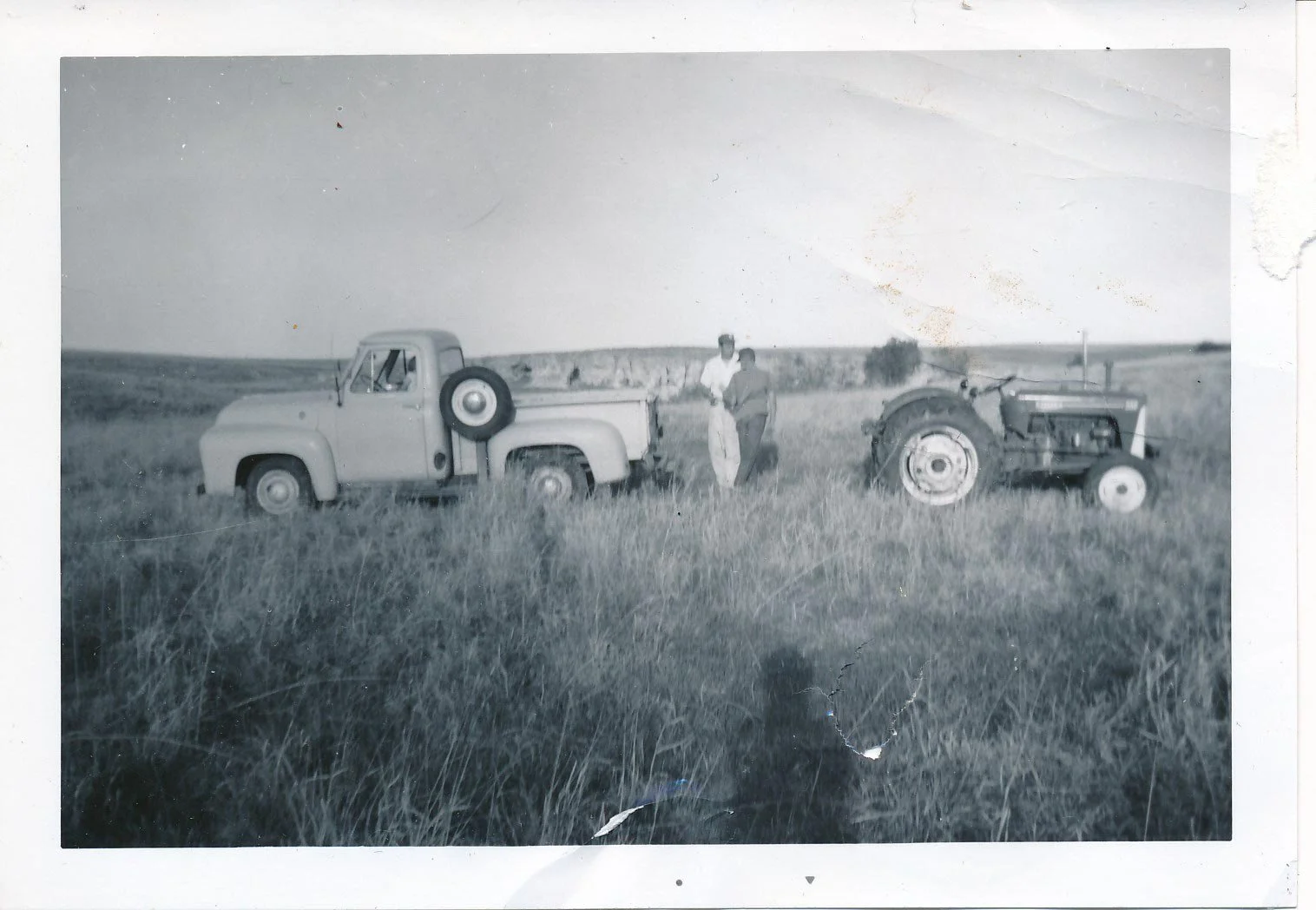 Black and white photograph of two men standing between two vintage tractors in a field with tall grass, with a distant landscape of hills and open sky.