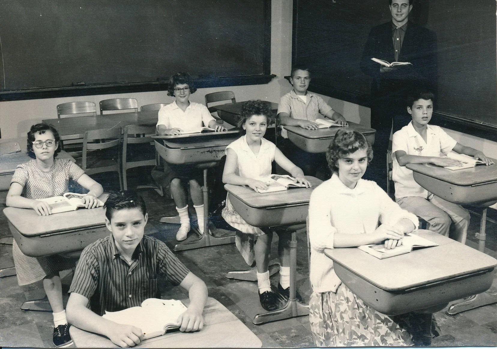 Black and white photo of a classroom with eight children seated at desks, each with an open book, and a teacher standing at the blackboard holding a book.