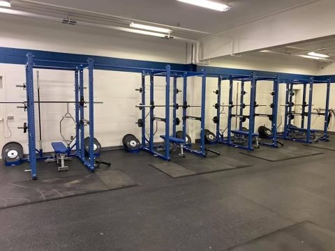 Empty gym with blue power racks, barbells, weight plates, and benches on rubber flooring.