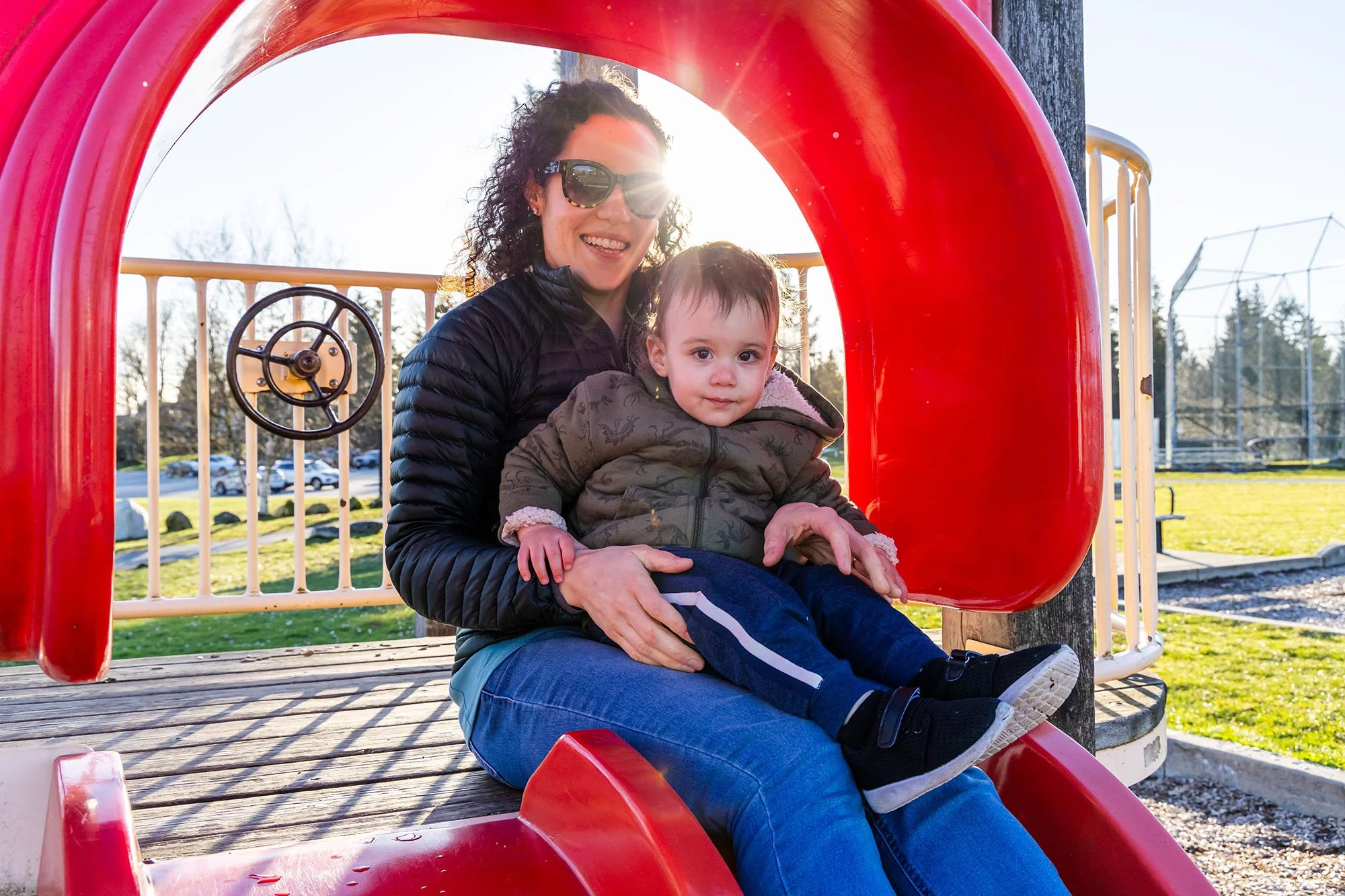 A woman and a young boy sitting on a red playground slide at a park, smiling with sunlight shining behind them.