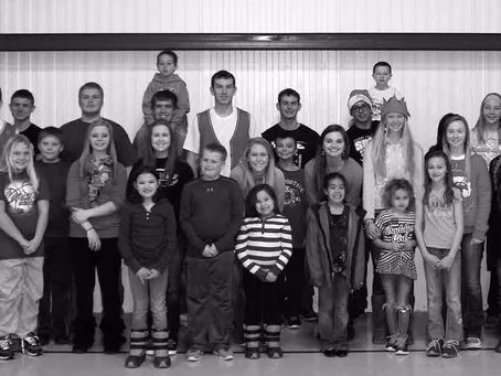 Group photo of children and teenagers in a school or classroom setting, some wearing casual clothes and smiling.