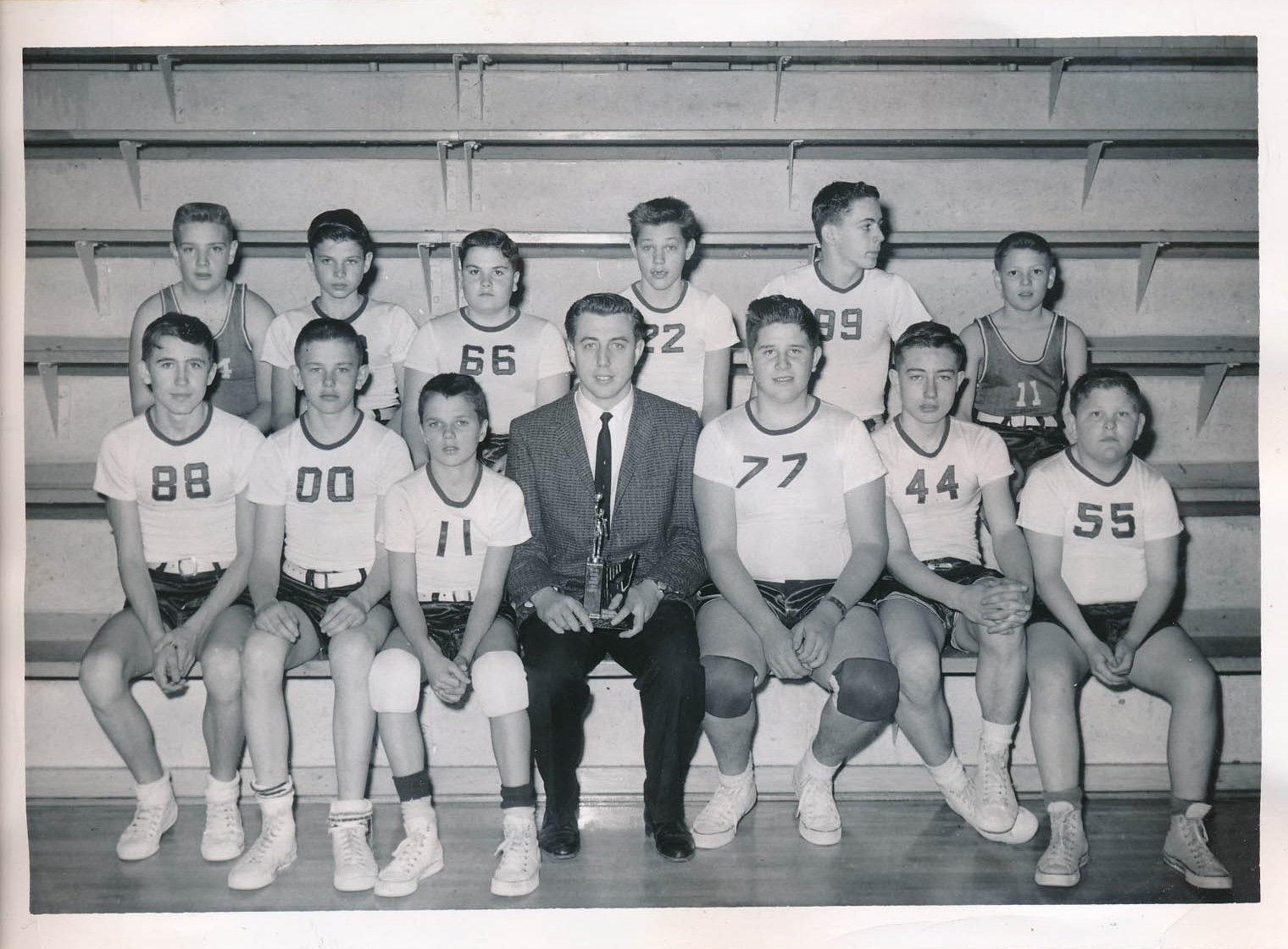 Black and white photo of a boys' basketball team with their coach, sitting and standing on gym bleachers. The team wears jerseys with numbers, shorts, and sneakers.