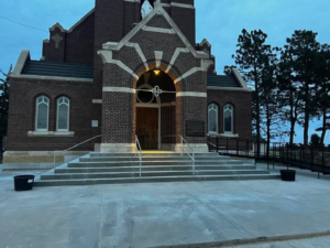 A Catholic church with brick exterior and a triangular roof, illuminated by a yellow light, situated on a hill with stairs leading up to the entrance, surrounded by pine trees at dusk.