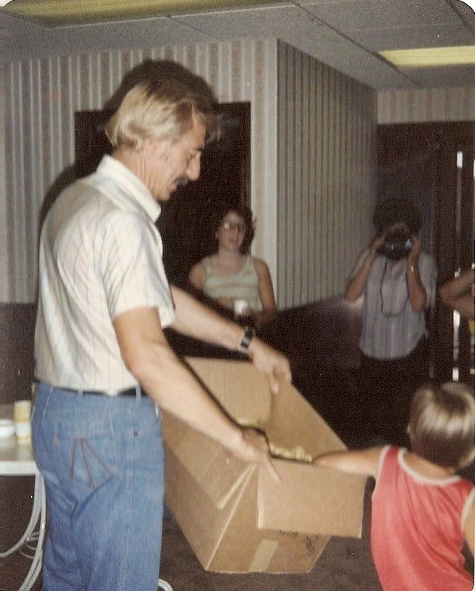 A man in a light-colored striped shirt and jeans is giving a gift to a young boy wearing a red sleeveless shirt, with two women observing in the background.