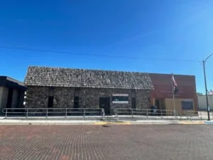 Empty brick building with a shingled roof and an American flag outside, along a brick sidewalk