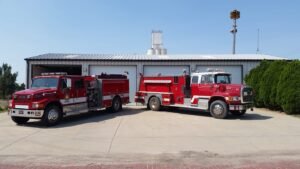 Two red fire trucks parked in front of a building with a hedge on the right side.