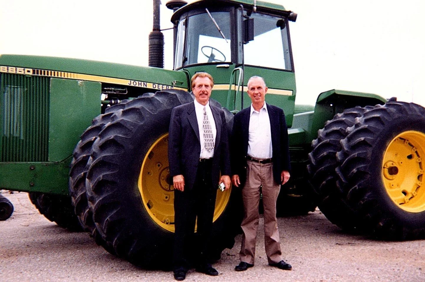 Two men standing in front of a large green John Deere tractor with yellow wheels, both wearing suits and smiling.