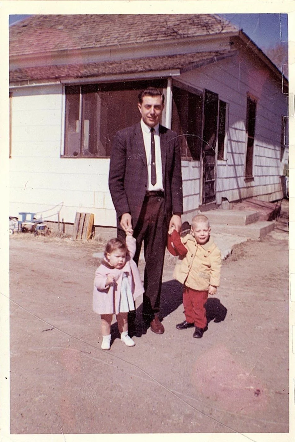 A man in a suit standing outside a house, holding hands with a young girl and a boy, both children in warm clothing.