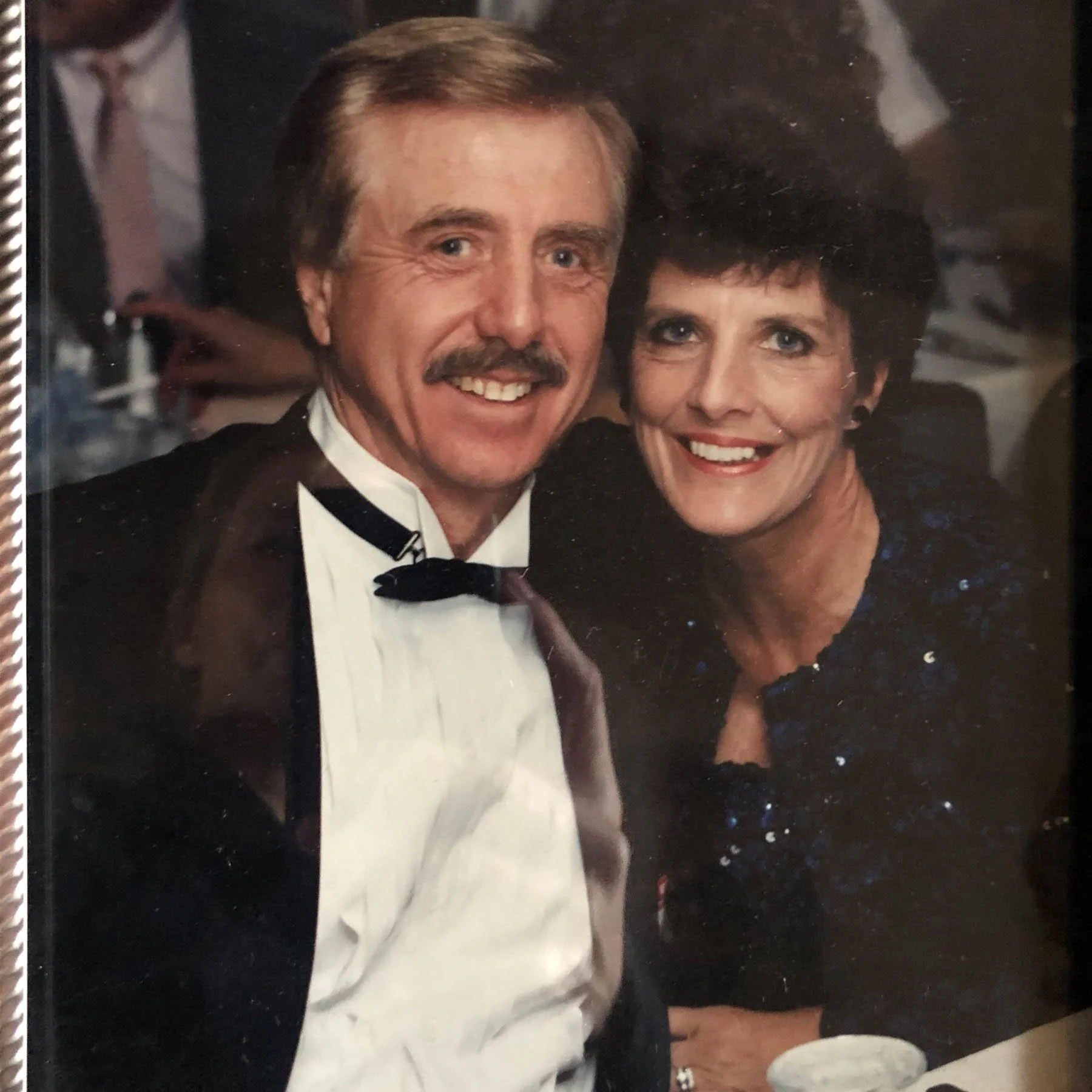 A smiling man with a mustache and light-colored hair in a tuxedo, sitting close to a woman with short dark hair and a sparkly dark dress, both at a formal event.