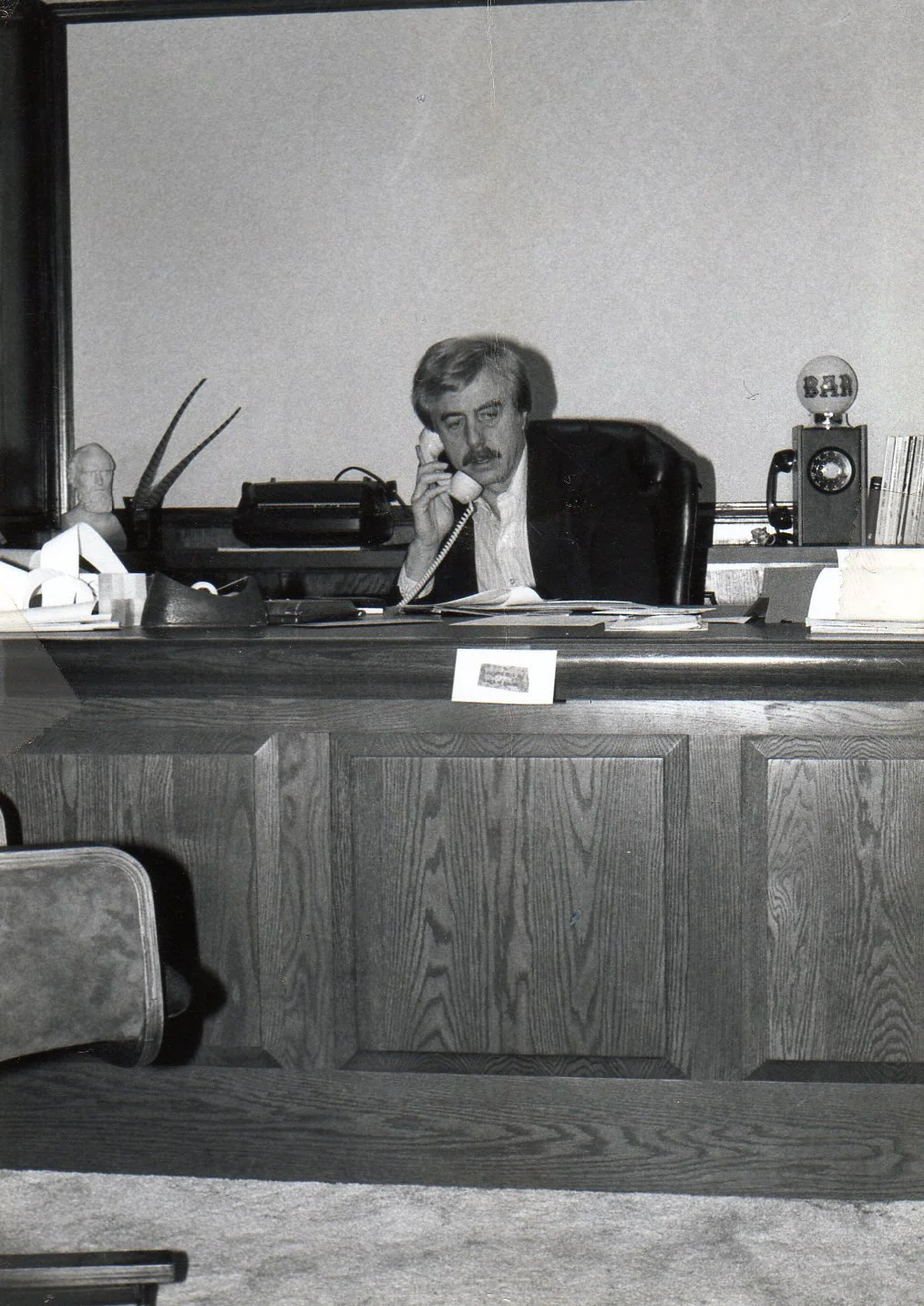 Black and white photo of a man with a mustache sitting at a desk, talking on a landline phone, in an office setting.