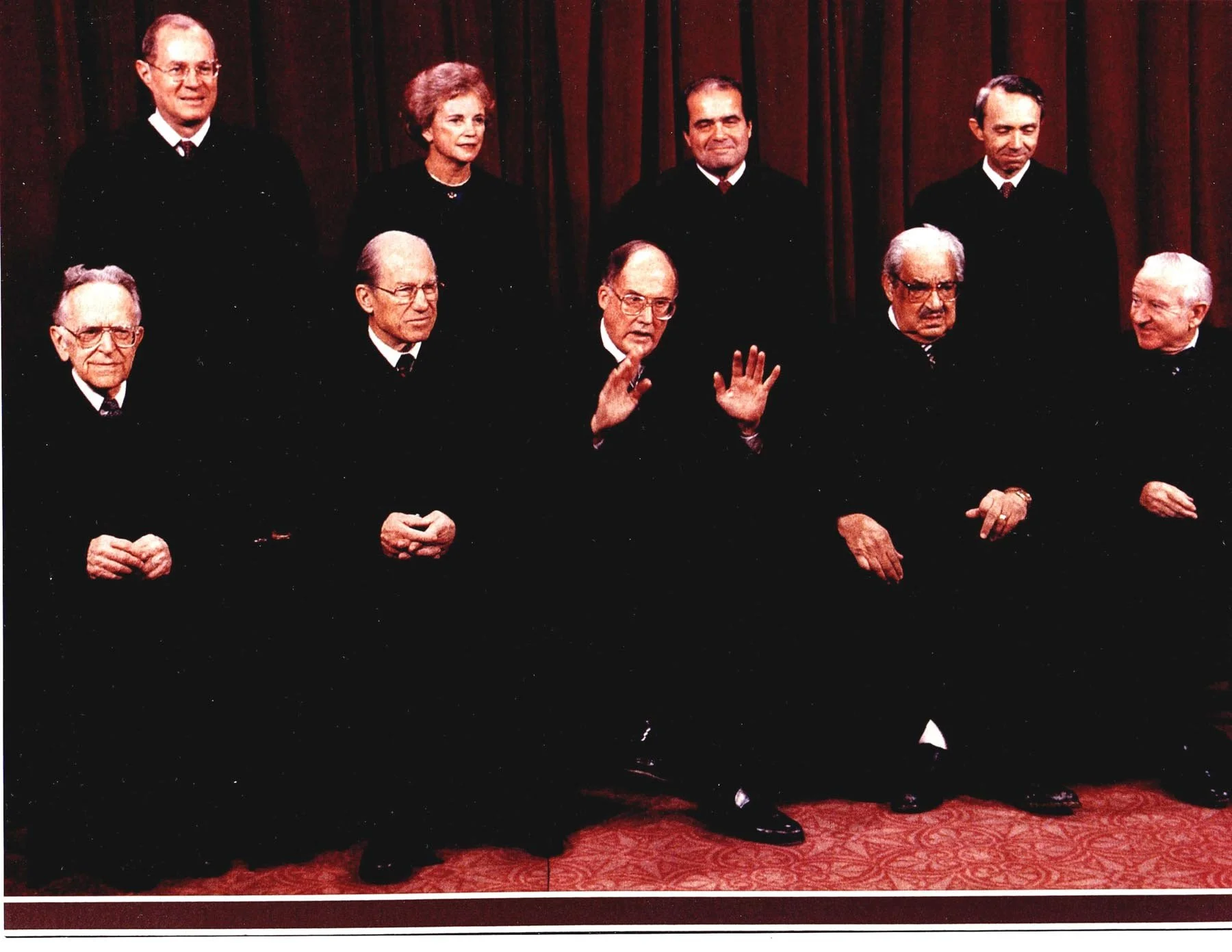 A group of nine Supreme Court justices posing for a formal photo in front of a red curtain. Some are seated, others standing, with Samuel Alito in the center raising his hands.