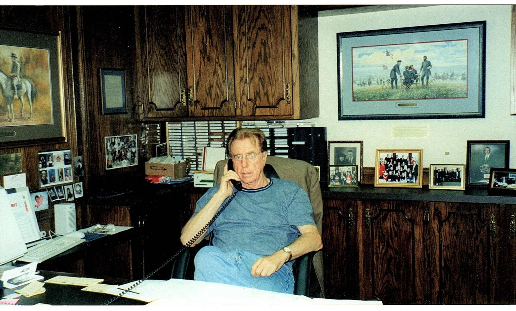 Man sitting at an office desk, talking on a landline phone, with framed pictures and artwork on the wood-paneled walls behind him.