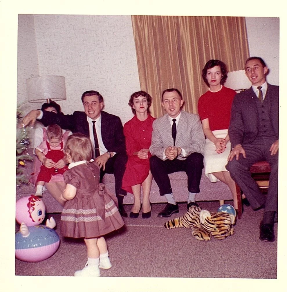 A vintage family photo from the 1950s or 1960s shows seven people, including three children, sitting on a couch in a living room. There is a decorated Christmas tree with pink and blue ornaments on the left, and a small stuffed tiger on the floor. Tw