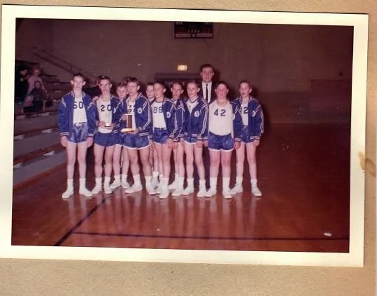 A group of young female athletes in matching athletic uniforms, standing on an indoor gym floor with a coach, holding a trophy.