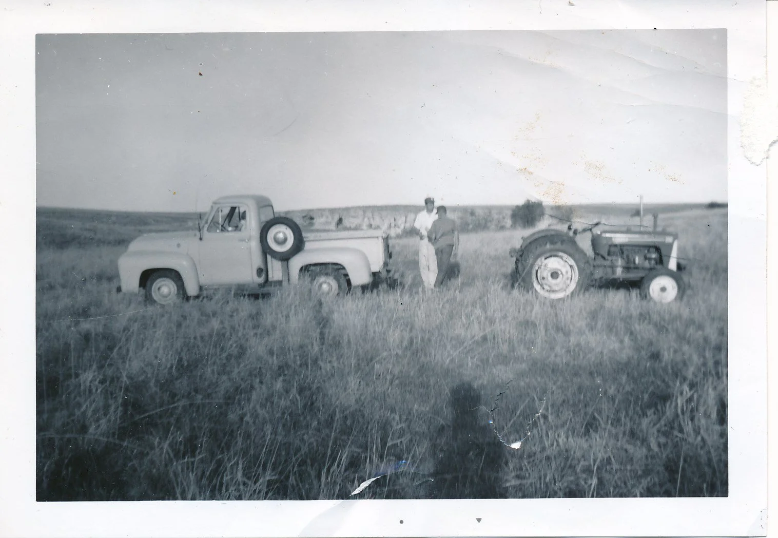 Black and white photo of a pickup truck and a tractor in a rural field with two men standing between them, farmland and a few trees in the background.