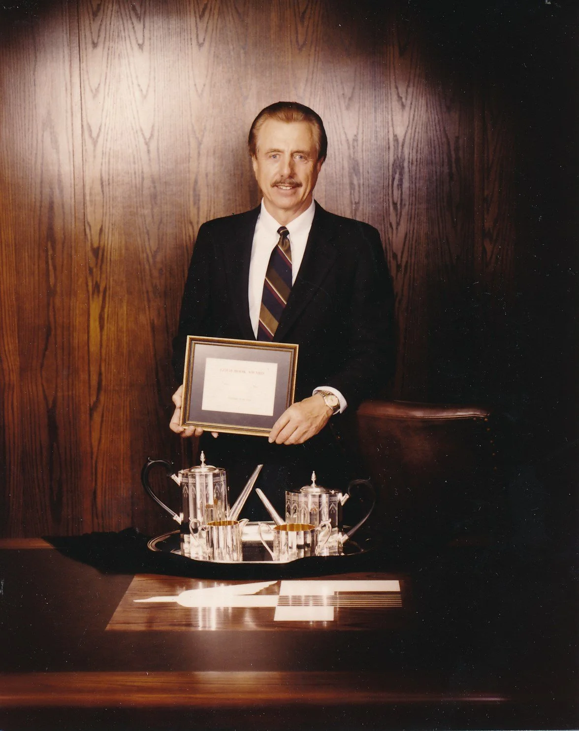 A man in a suit holding a framed certificate, standing behind a table with a tea set and documents.