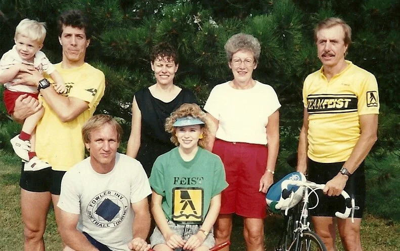 A group of seven people, including children and adults, posing outdoors in front of trees. Some are smiling, and there is a bicycle on the right.