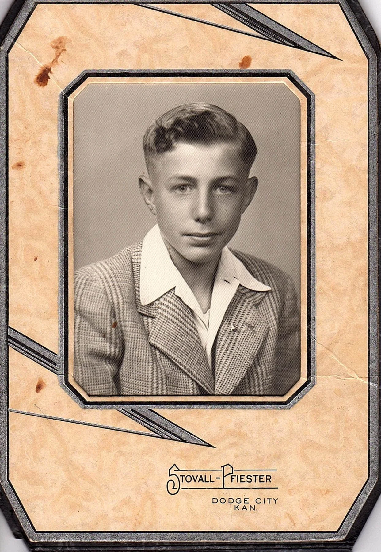 Vintage portrait photograph of a young man with short hair, wearing a checkered blazer and a white collared shirt, framed on an aged paper background with decorative lines.