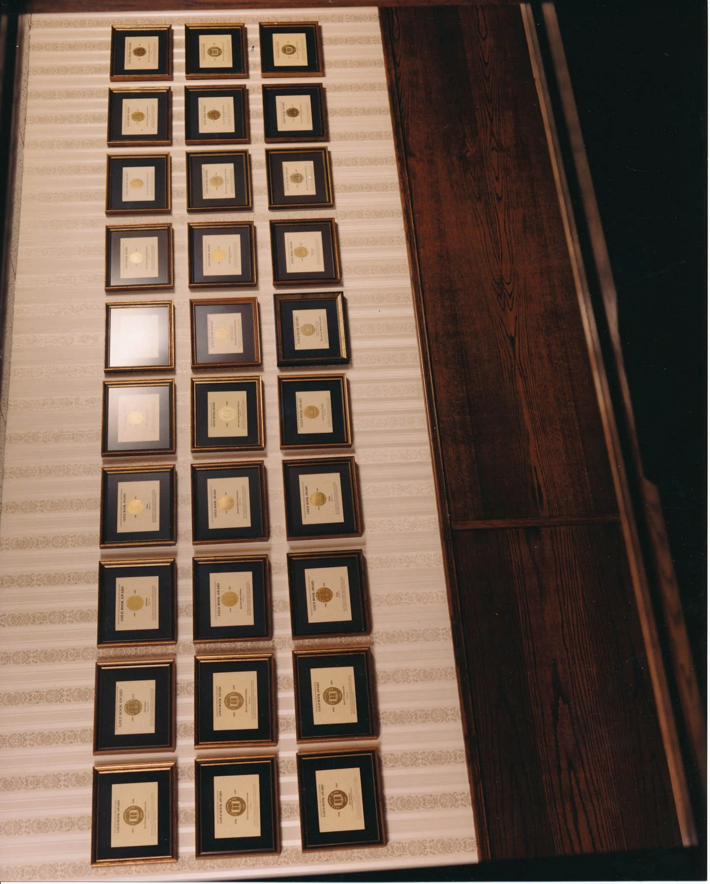Wall with 30 framed certificates or awards arranged in five rows and six columns, next to a dark wooden panel.