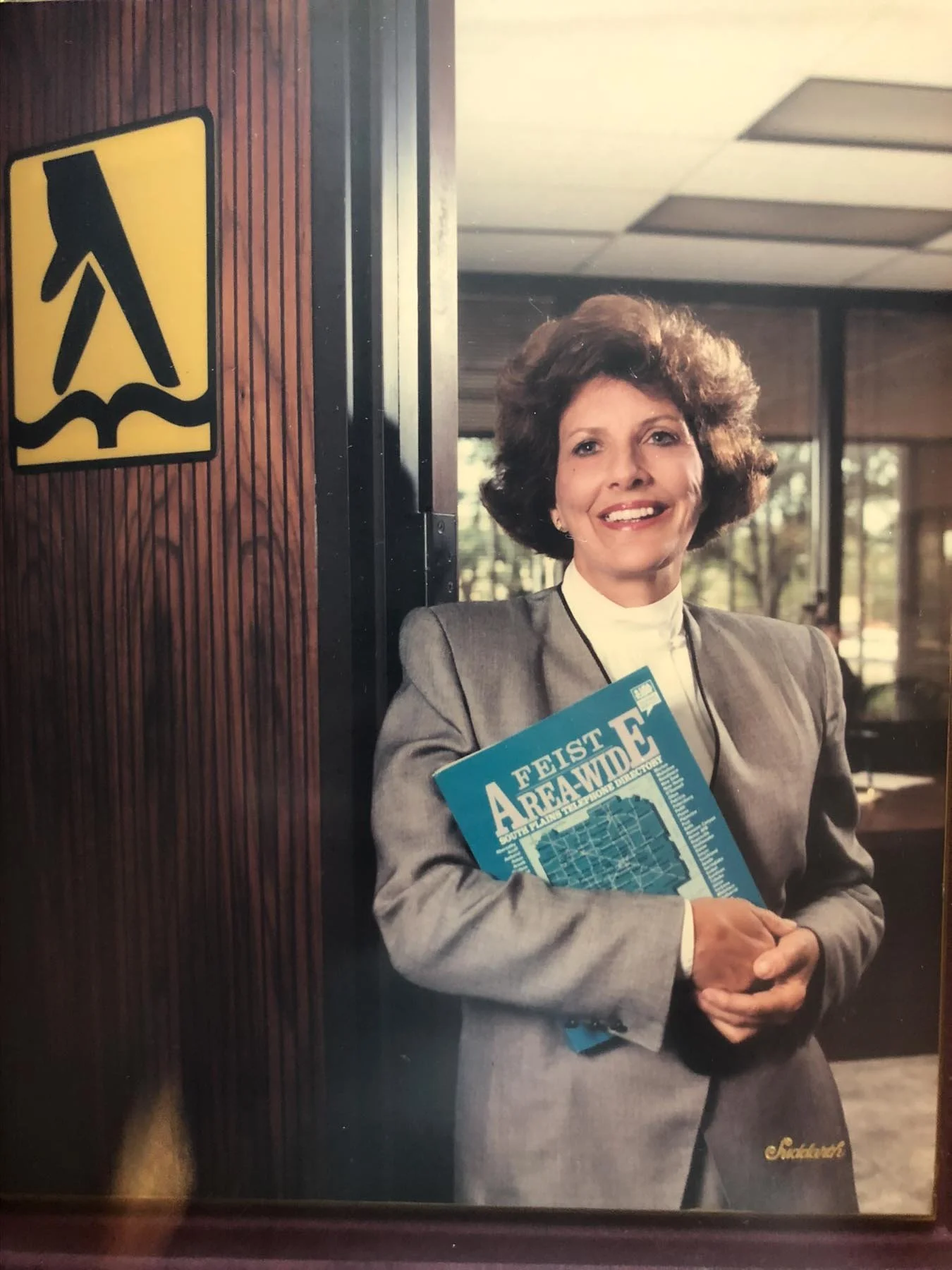 A woman with curly hair wearing a gray suit and white shirt, smiling and holding a blue blueprint titled 'FEIST AREA WIDE' inside an office or conference room.