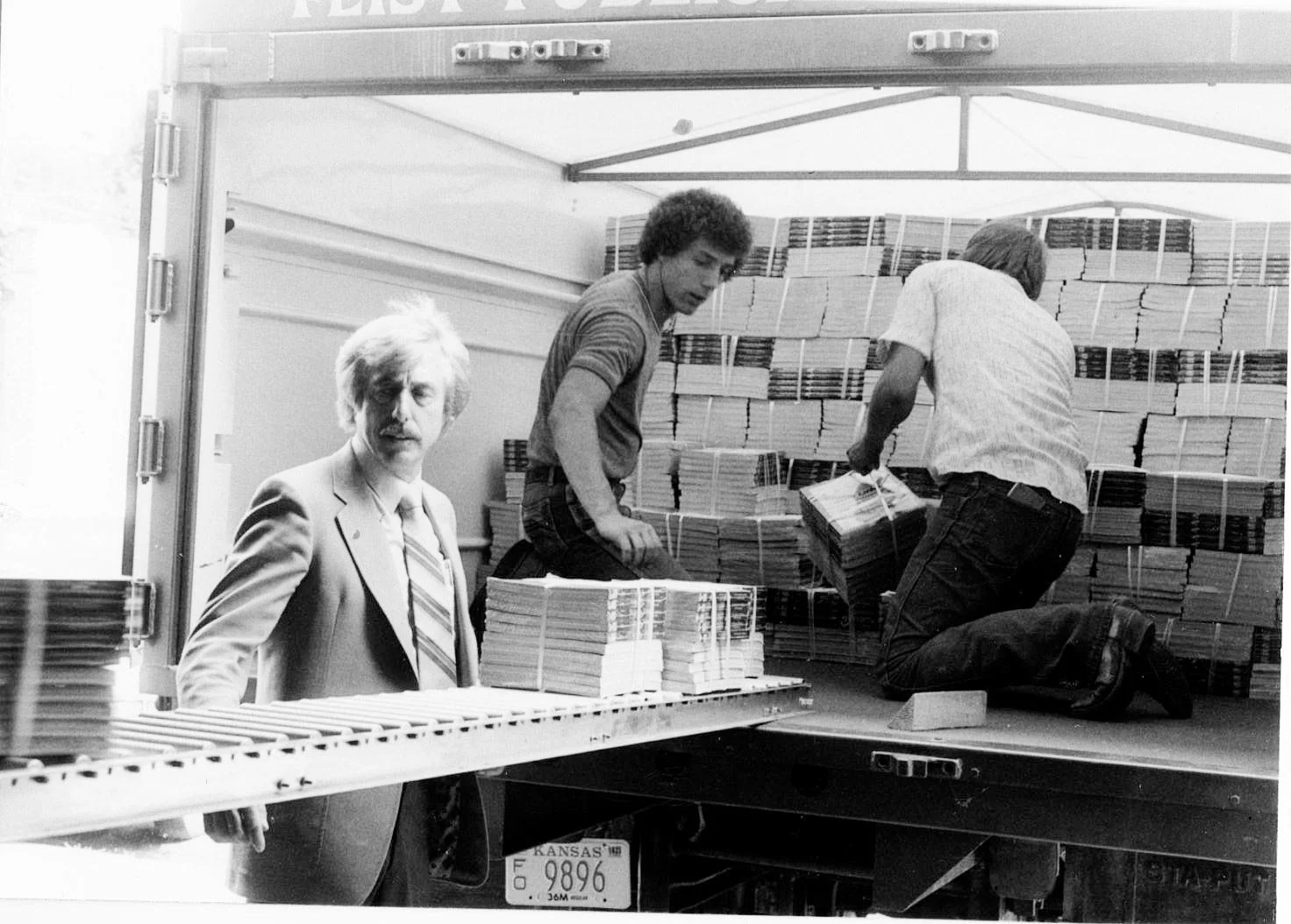 Black and white photo of three men packing books into the back of a truck. One man in a suit stands near the truck, while two men inside the truck are stacking and handling books.