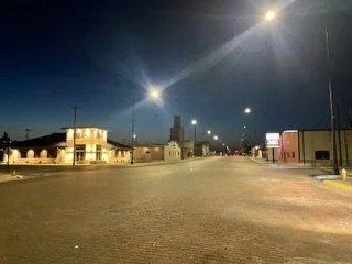 Empty street at night with streetlights illuminating the area and a few buildings in the background.