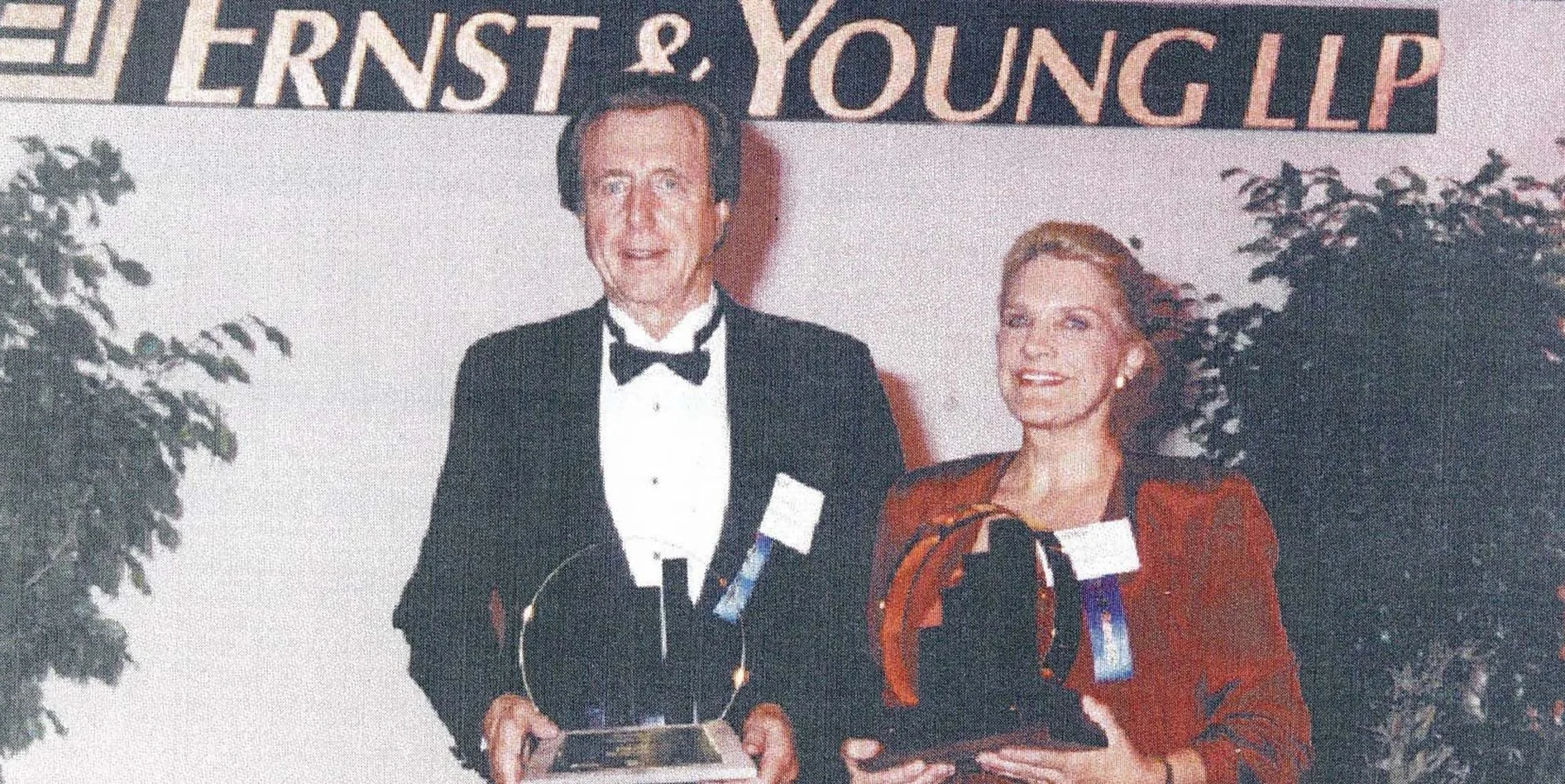 A man and a woman dressed in formal attire standing together at an awards event, holding plaques and smiling at the camera, with a banner above them reading from left to right: 'ERNST' and 'YOUNG LP'.