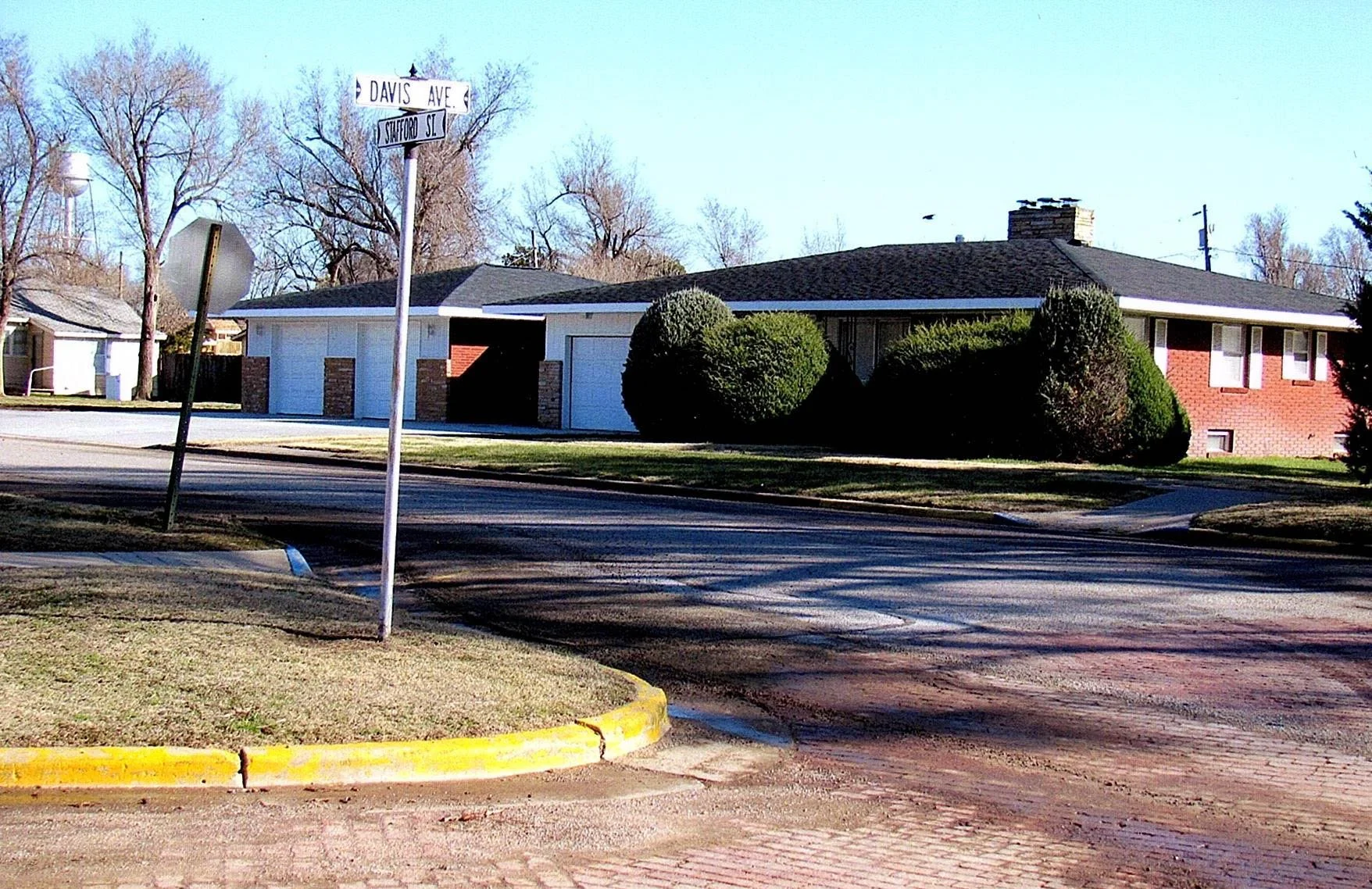 Residential street corner with a house, trees, street signs reading Davis Ave and Stafford St, and a traffic sign leaning to the side.