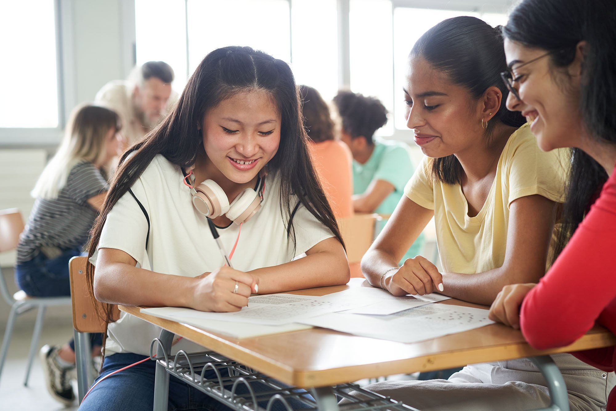 Three students sitting at a desk in a classroom, working together on a paper, smiling and engaging in discussion.