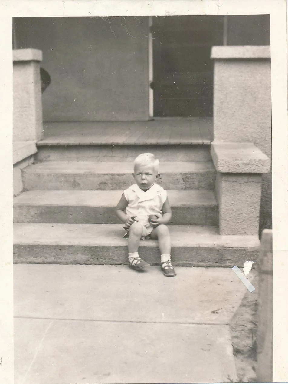 A child sitting on the steps of a porch, looking at the camera, in a black and white photo.