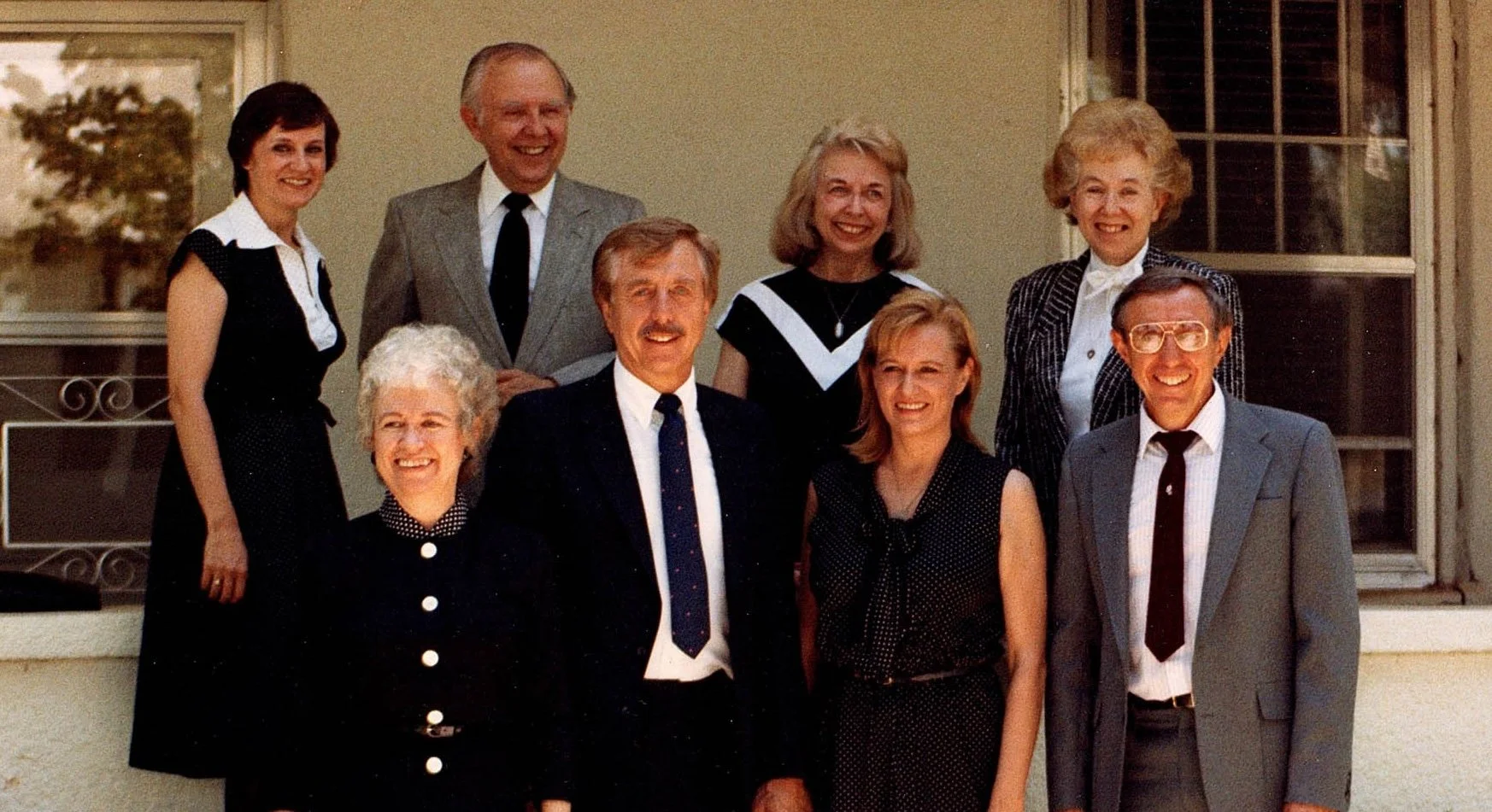 Group of nine adults posing on a porch, dressed in vintage clothing, smiling at the camera.
