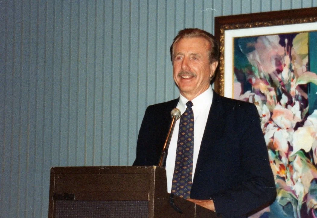 A man in a suit and tie stands at a podium with a microphone, speaking or giving a presentation with a smile, in front of a blue wall with vertical paneling and an abstract colorful painting.