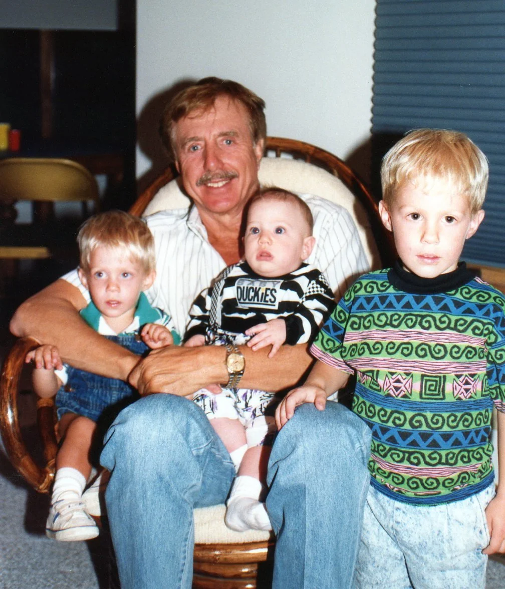 An older man with a mustache sitting in a wooden chair holding three young boys on his lap and beside him.