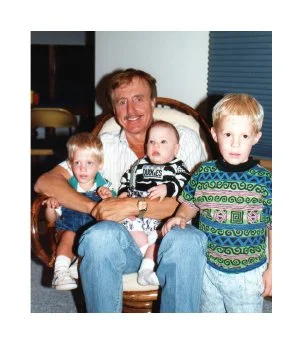 An older man sitting on a wooden chair with three children around him, in an indoor setting.