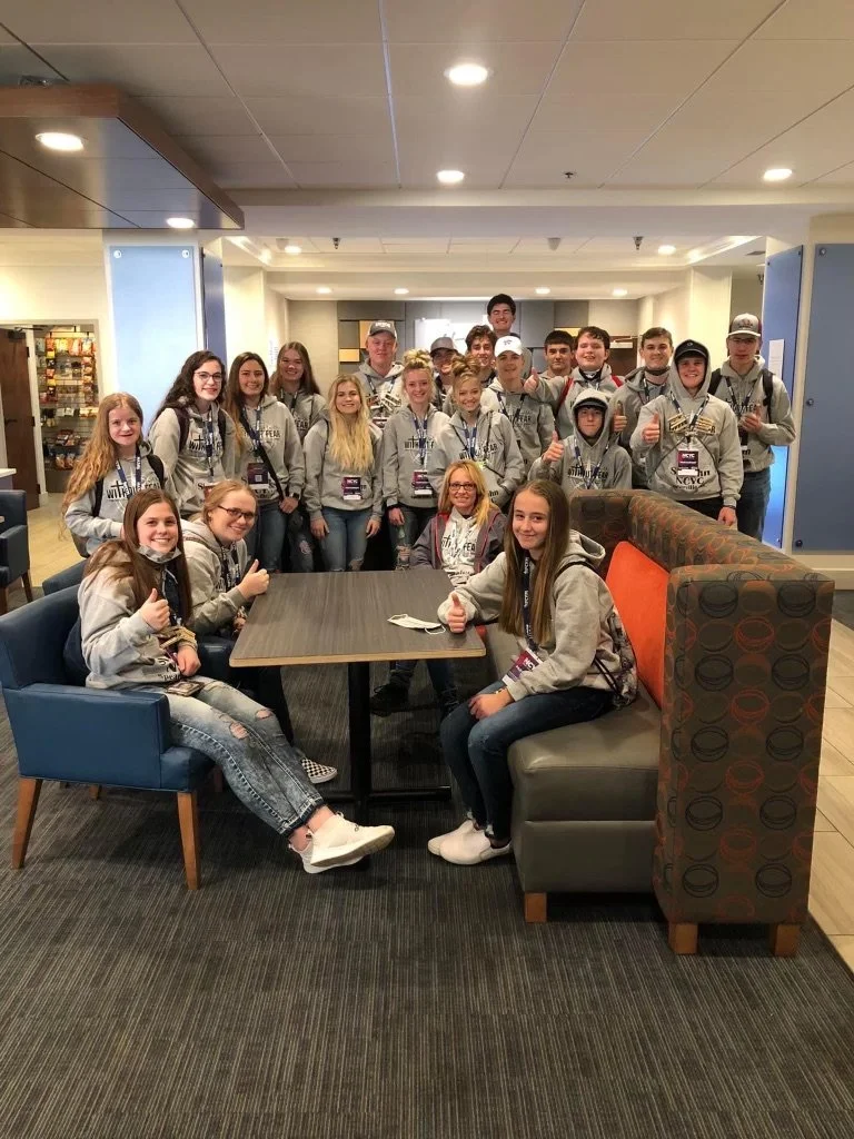 Group of teenagers and young adults posing for a photo in a hotel lobby, some wearing matching gray hoodies and conference badges, smiling and giving thumbs-up.