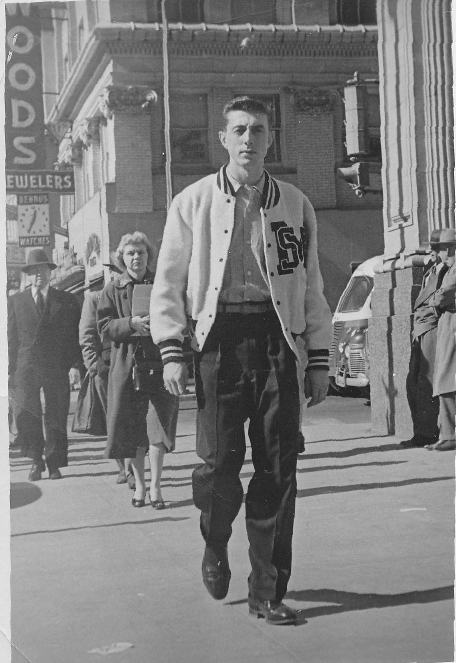 A young man in a varsity jacket walking on a city street, with other pedestrians and a vintage vehicle in the background.