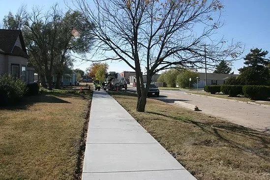 Sidewalk in a suburban neighborhood with houses and trees, some with green leaves, and a few cars parked on the street.