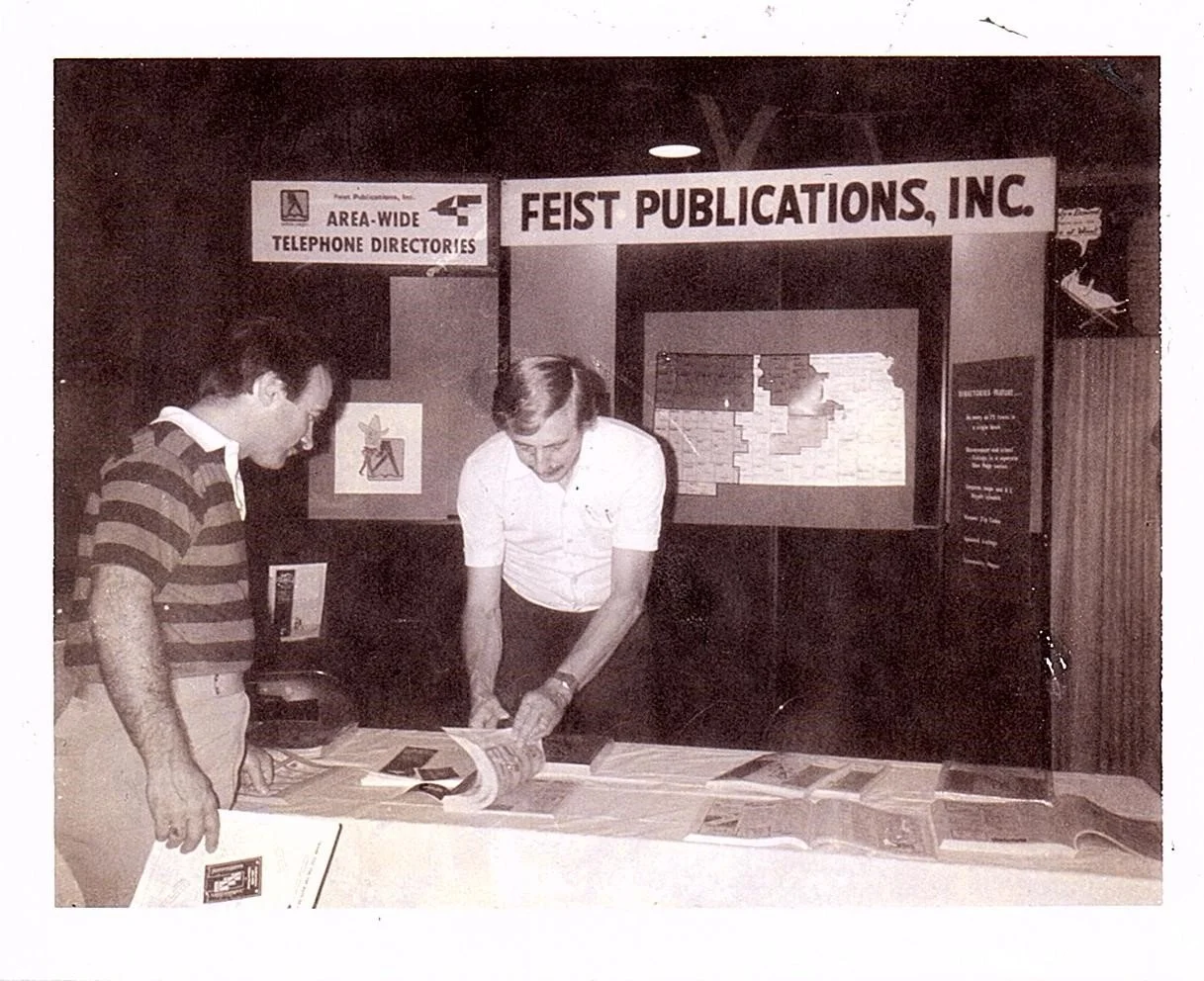 Two men looking at maps and publications on a table at a booth for FEIST PUBLICATIONS, INC. at an event, with signs indicating area-wide telephone directories and a map display in the background.