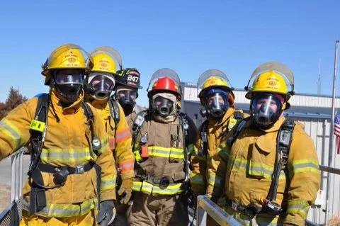 Six firefighters in yellow gear and helmets, with one in the middle wearing a tan uniform and red helmet, standing outdoors on a sunny day.