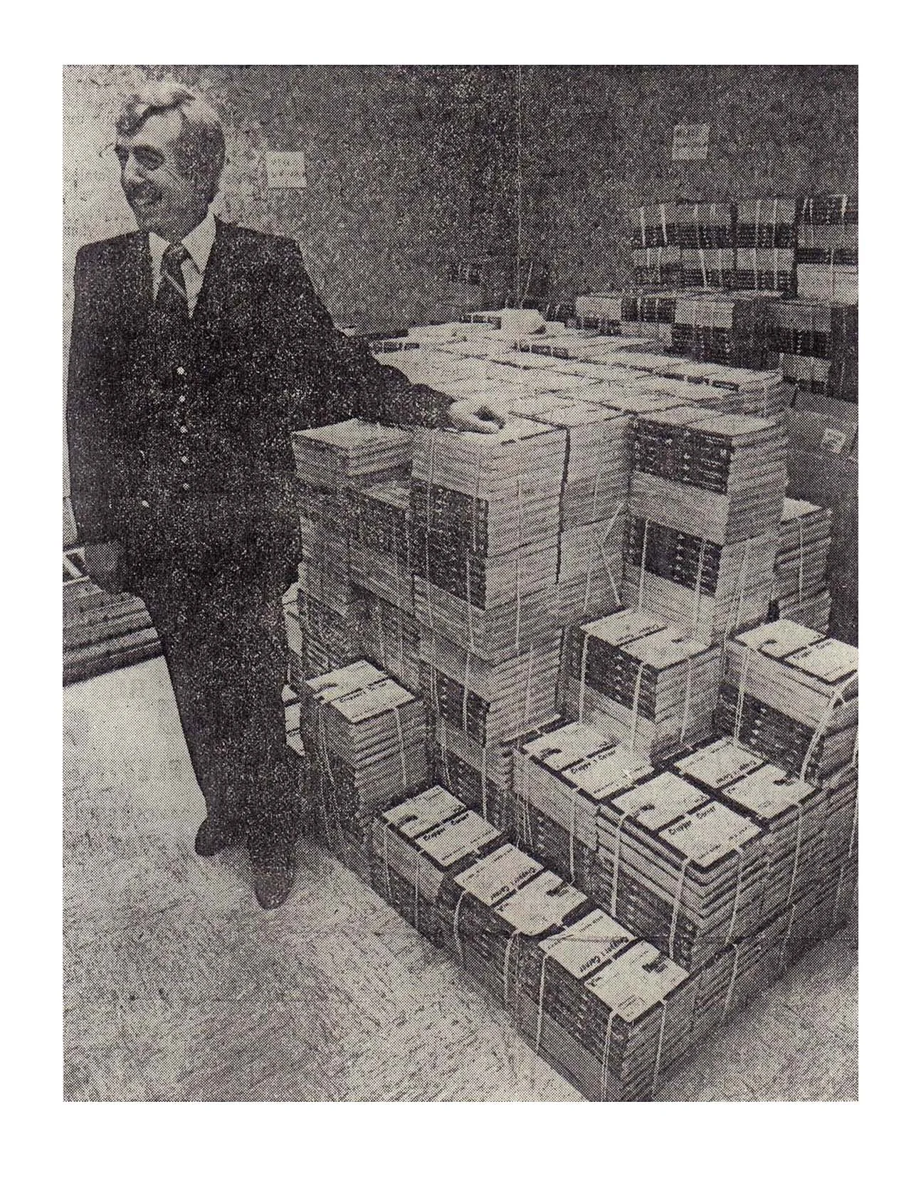 A man in a suit standing beside stacks of newspapers or magazines in an office or warehouse setting.