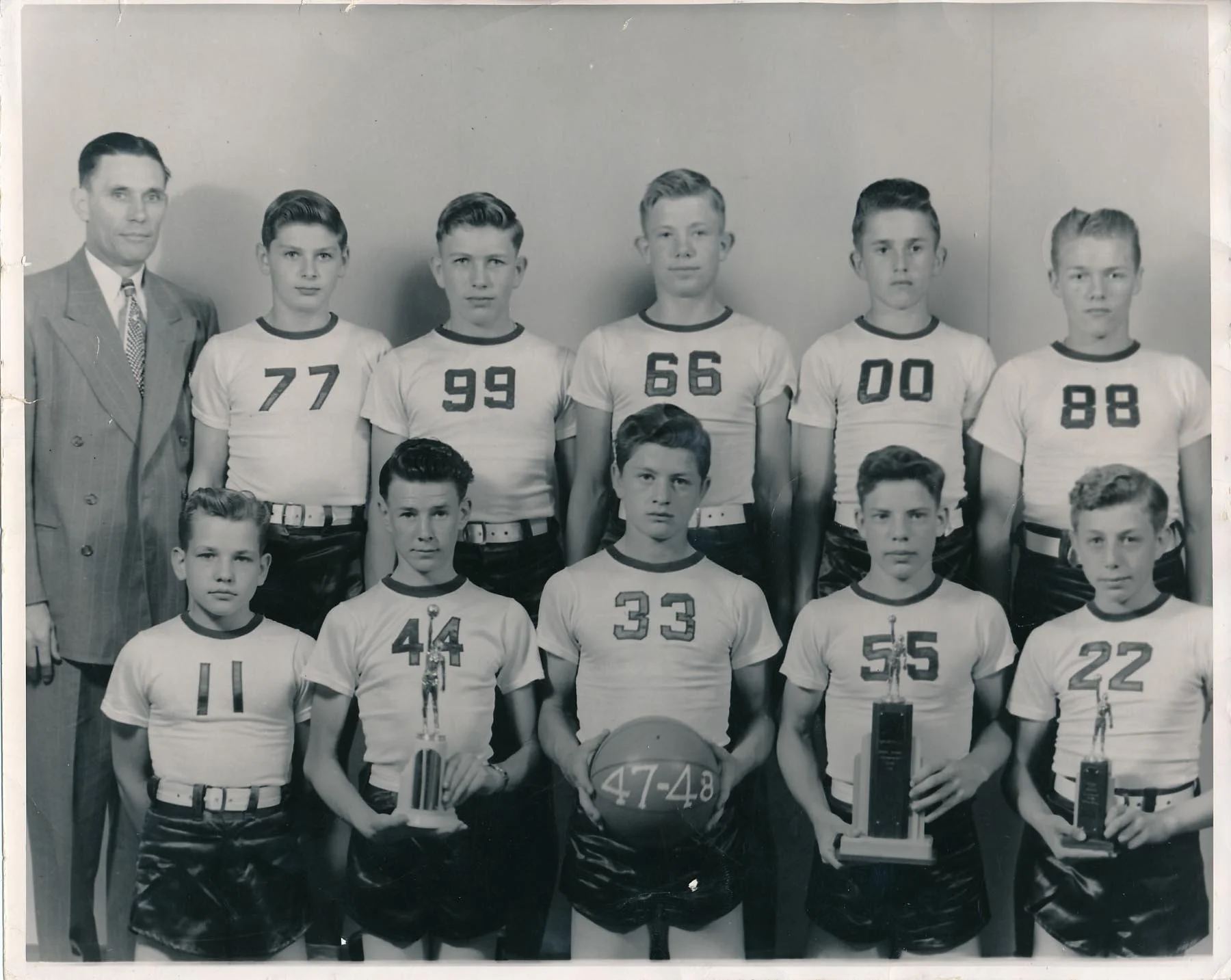 A black and white team photograph of boys in sports uniforms with numbers, along with one adult man in a suit, posing against a plain wall. The boys are holding trophies and a basketball.