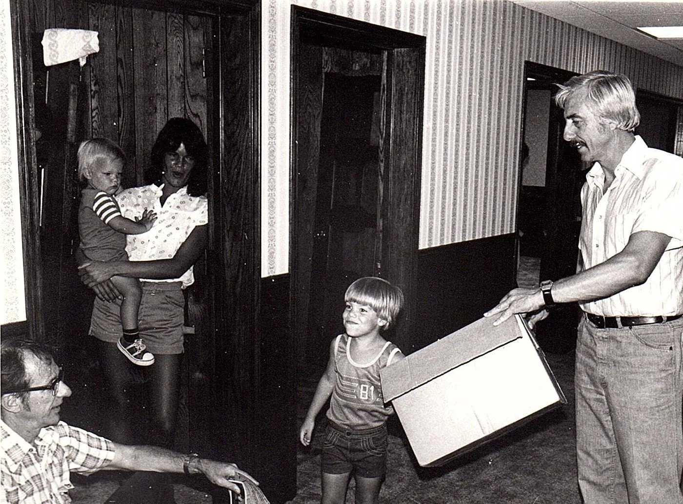 A man with gray hair and wearing a short-sleeve button-up shirt is giving a box to a young boy with blonde hair and a striped tank top, inside a room with wood-paneled walls. A woman holding a young girl in a striped shirt is standing nearby, and an 