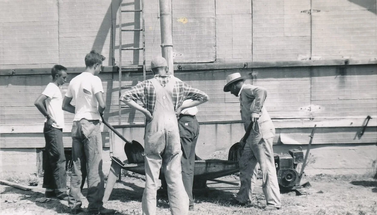 Five people working together outdoors, with some using shovels and a wheelbarrow, near a brick building.