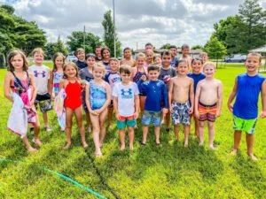 Group of children and teenagers in swimsuits standing on grassy field at outdoor swimming pool area