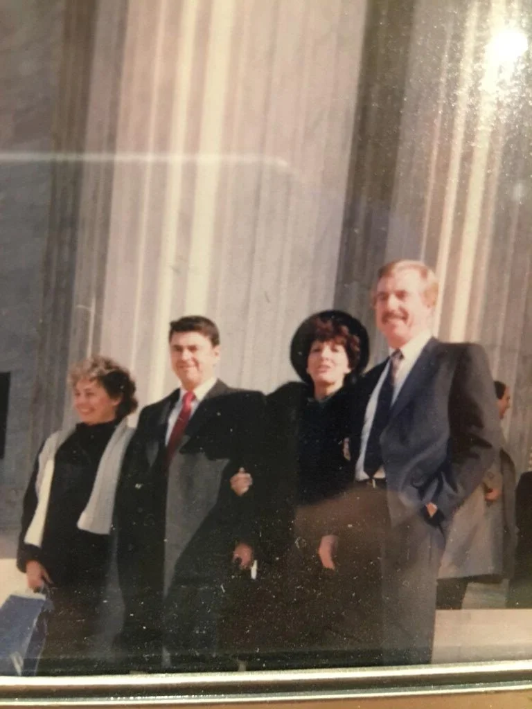 Four people, three women and one man, standing together inside a large building with tall columns, dressed in formal attire, smiling for the photo.